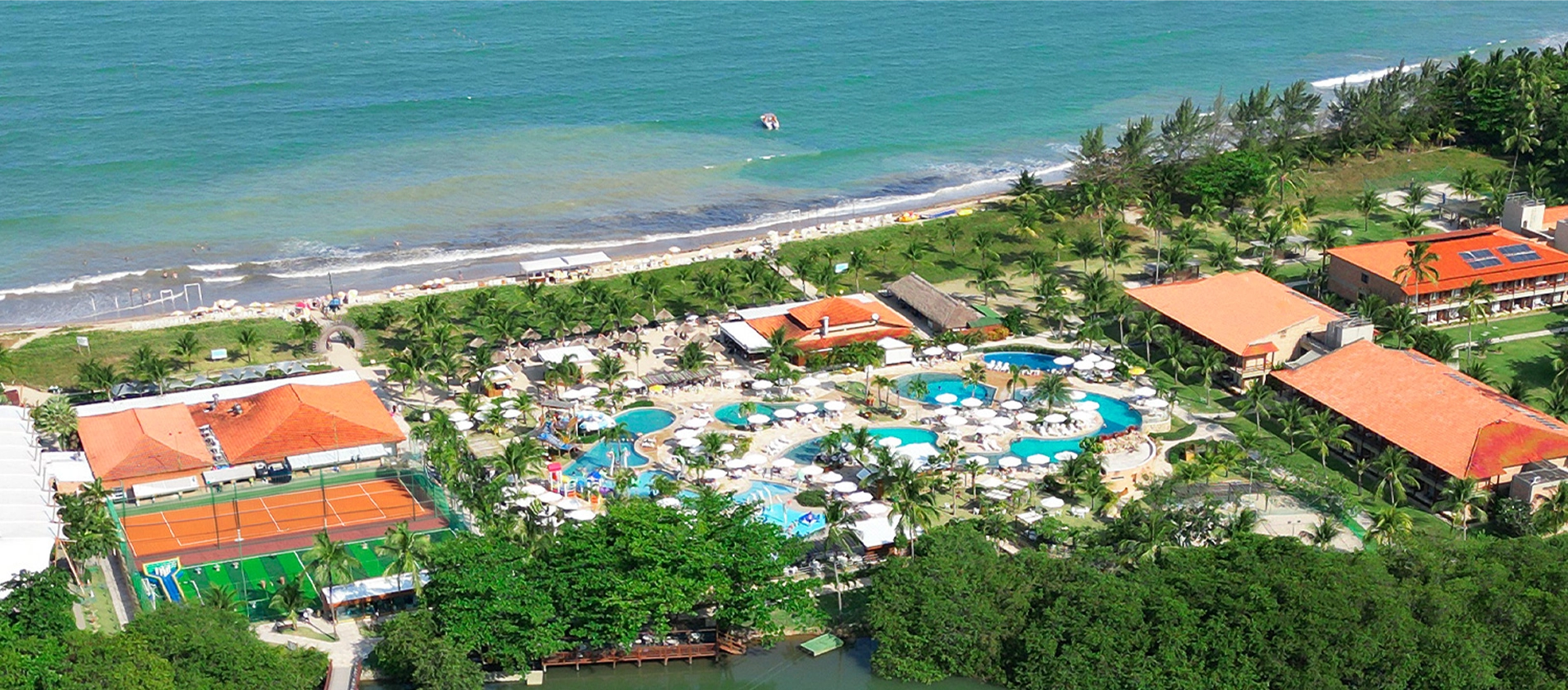 Imagem aérea do Salinas Maragogi mostrando o complexo de piscinas, dos quartos, pontos de consumo e ao fundo a imensidão da praia. Todo o resort é cercado por área verde e o um rio passa pelo meio do resort.