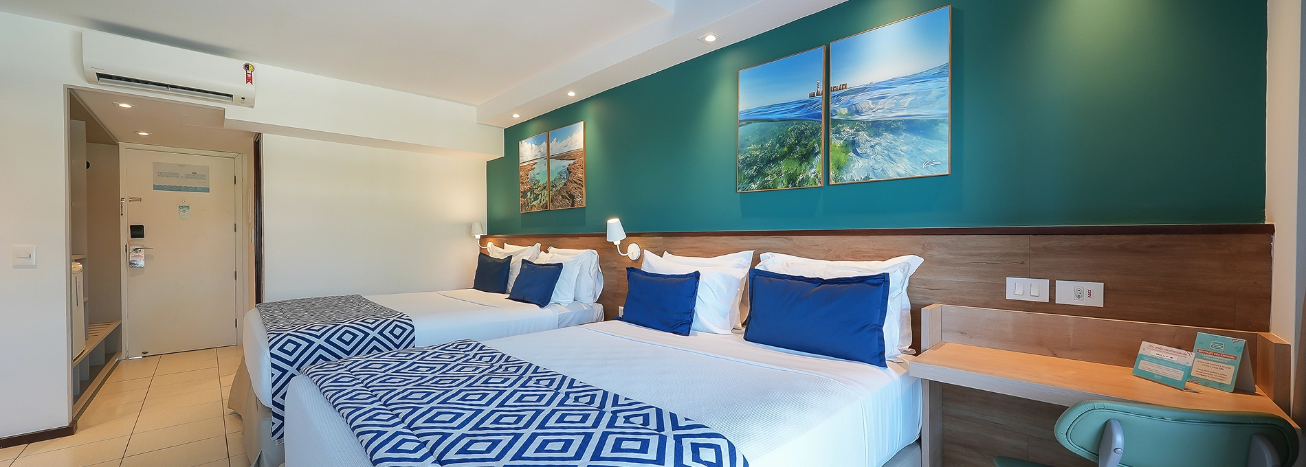 Visão interna da Suíte Familiar. Duas camas queen size, duas luminárias, sendo uma para cada cama, uma cabeceira de madeira. Uma escrivaninha com cadeira verde ao lado da cama do lado direito. Parede com cor verde turquesa com quadros pendurados, dois em cada topo da cama. Ao fundo a porta de saída do quarto e em cima, uma pequena visão do mezanino.