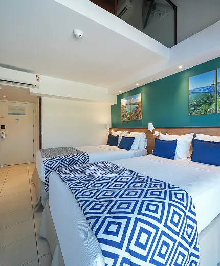 Visão interna da Suíte Familiar. Duas camas queen size, duas luminárias, sendo uma para cada cama, uma cabeceira de madeira. Uma escrivaninha com cadeira verde ao lado da cama do lado direito. Parede com cor verde turquesa com quadros pendurados, dois em cada topo da cama. Ao fundo a porta de saída do quarto e em cima, uma pequena visão do mezanino.