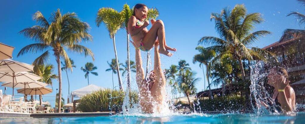 Criança feliz brincando na piscina sob um céu ensolarado com palmeiras ao fundo. Diversão e lazer em um dia de verão com amigos e família.