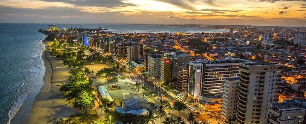 Vista panorâmica da orla de Maceió ao entardecer, com praia, prédios modernos e linhas de iluminação que destacam a elegância urbana.