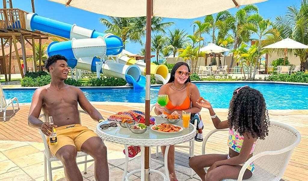 Família aproveitando dia ensolarado na piscina com área de diversão aquática, com adultos e uma criança sorrindo e confraternizando, ambiente relaxante e paradisíaco.