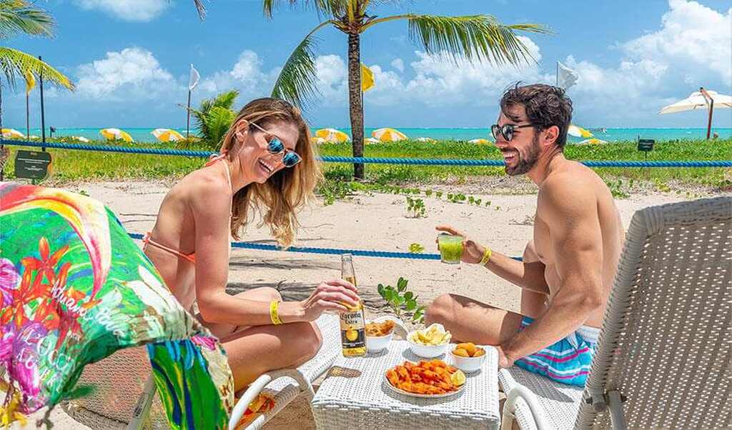 Casal feliz curtindo uma tarde na praia com drinks e petiscos sob o sol, com fundo de céu azul, mar e coqueiros.