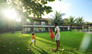 Família brincando de vôlei com bola em frentes a bloco de apartamento em fim do dia. Estão em um gramado bem verde.