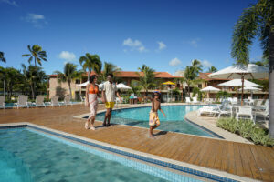 Família atravessando ponto do Salinas Maragogi em área de lazer, com piscinas ao redor, bloco de apartamentos ao fundo e céu sem nuvens.