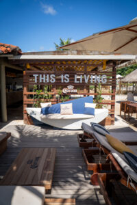 Banco com formato diferente, tem almofadas da corona azul e bege. Ao fundo um pallet de madeira sendo uma parede escrito "This is Living" e abaixo a logo da Corona. E mais a frente espreguiçadeiras e mesas.