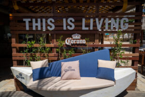Banco com formato diferente, tem almofadas da corona azul e bege. Ao fundo um pallet de madeira sendo uma parede escrito "This is Living" e abaixo a logo da Corona.