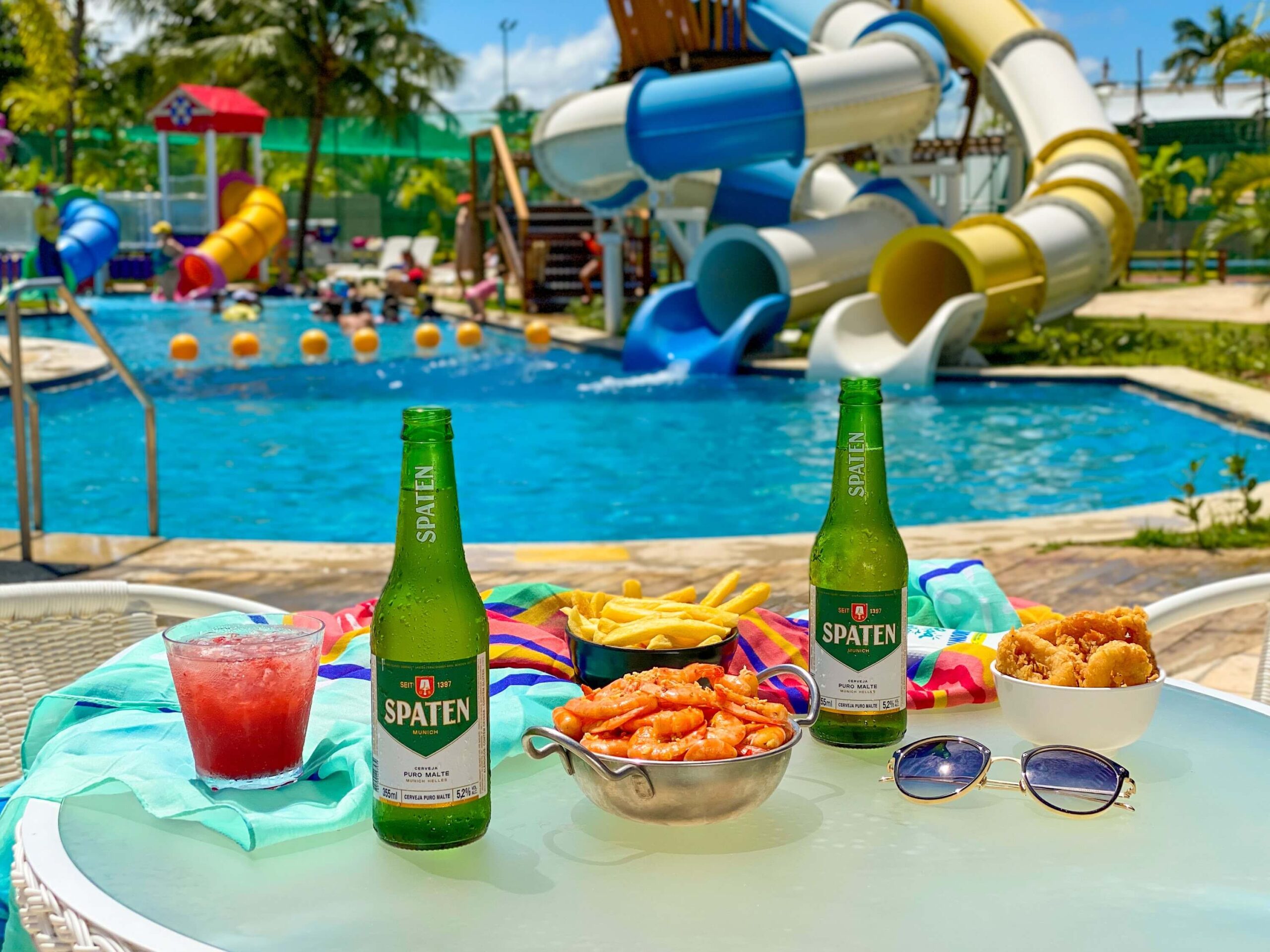 Duas long neck da cerveja Spaten ao lado de bowls de petiscos e de drink, servidos sobre a mesa, em frente à piscina infantil do Coral Bar, no resort Salinas Maragogi.