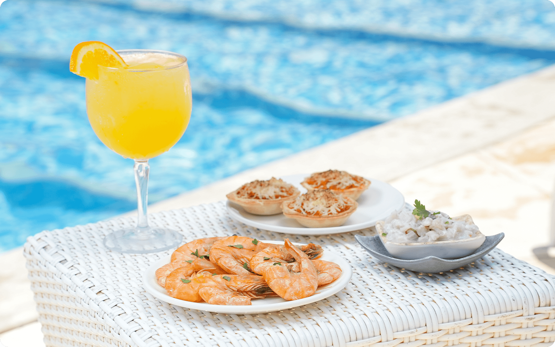 Petiscos como camarão, tortinha salgadas, com um drink amarelo expostos em um banco branco a beira da piscina em dia ensolarado.