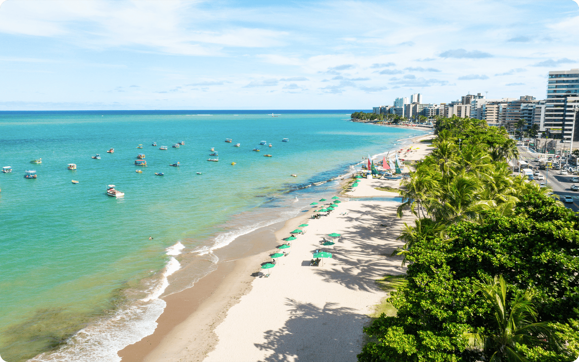 Vista áerea da praia de Jatiúca em Maceió. Com barcos coloridos na água, grande faixa de areia com guarda-sóis, vários coqueiros separando a praia da pista de carro. Com várias prédios ao fundo.