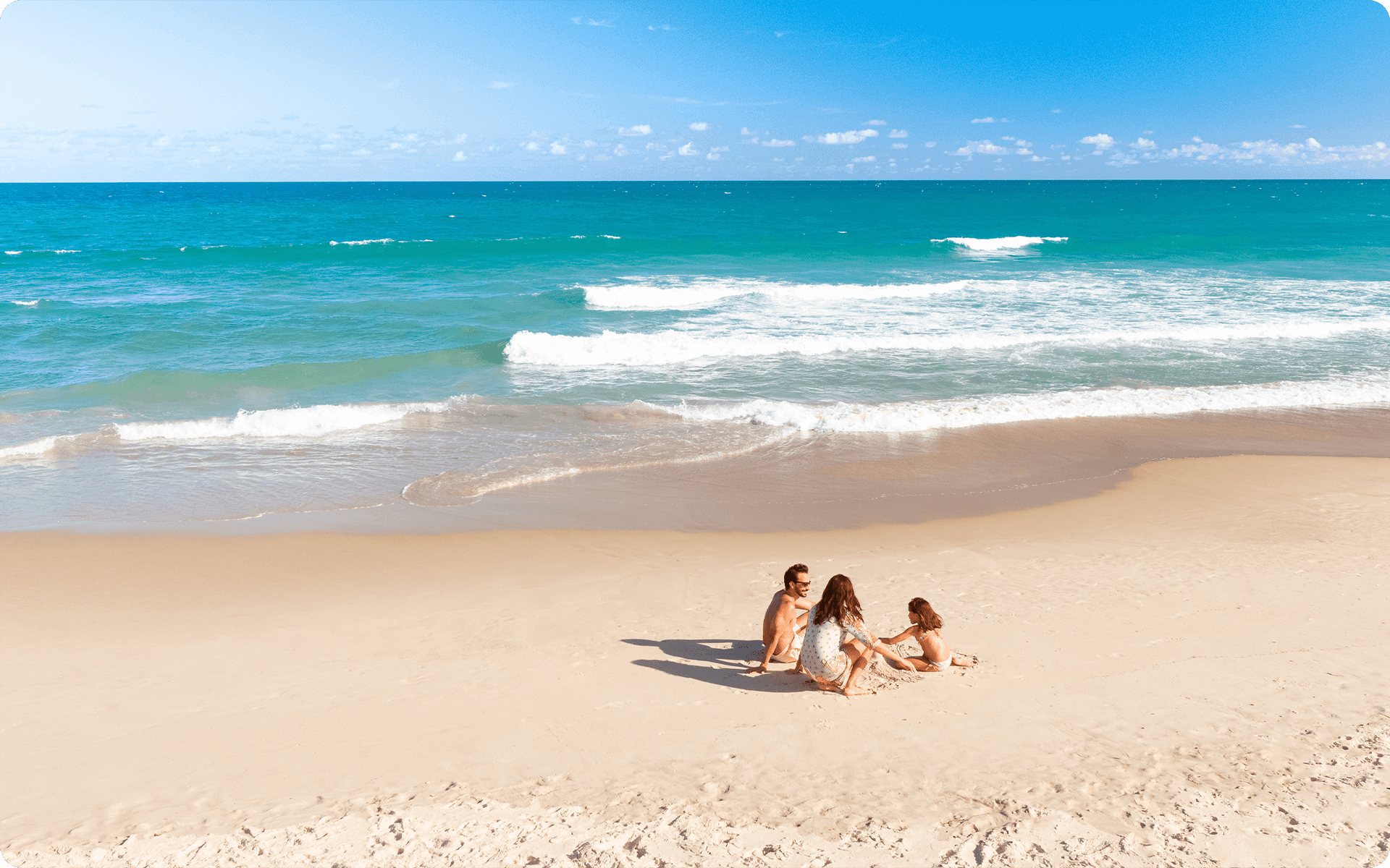Família sentada na areia, os pais estão brincando com a filha. Ao fundo há o mar com pequenas onda e seu azul é claro. O céu está com poucas nuvens.