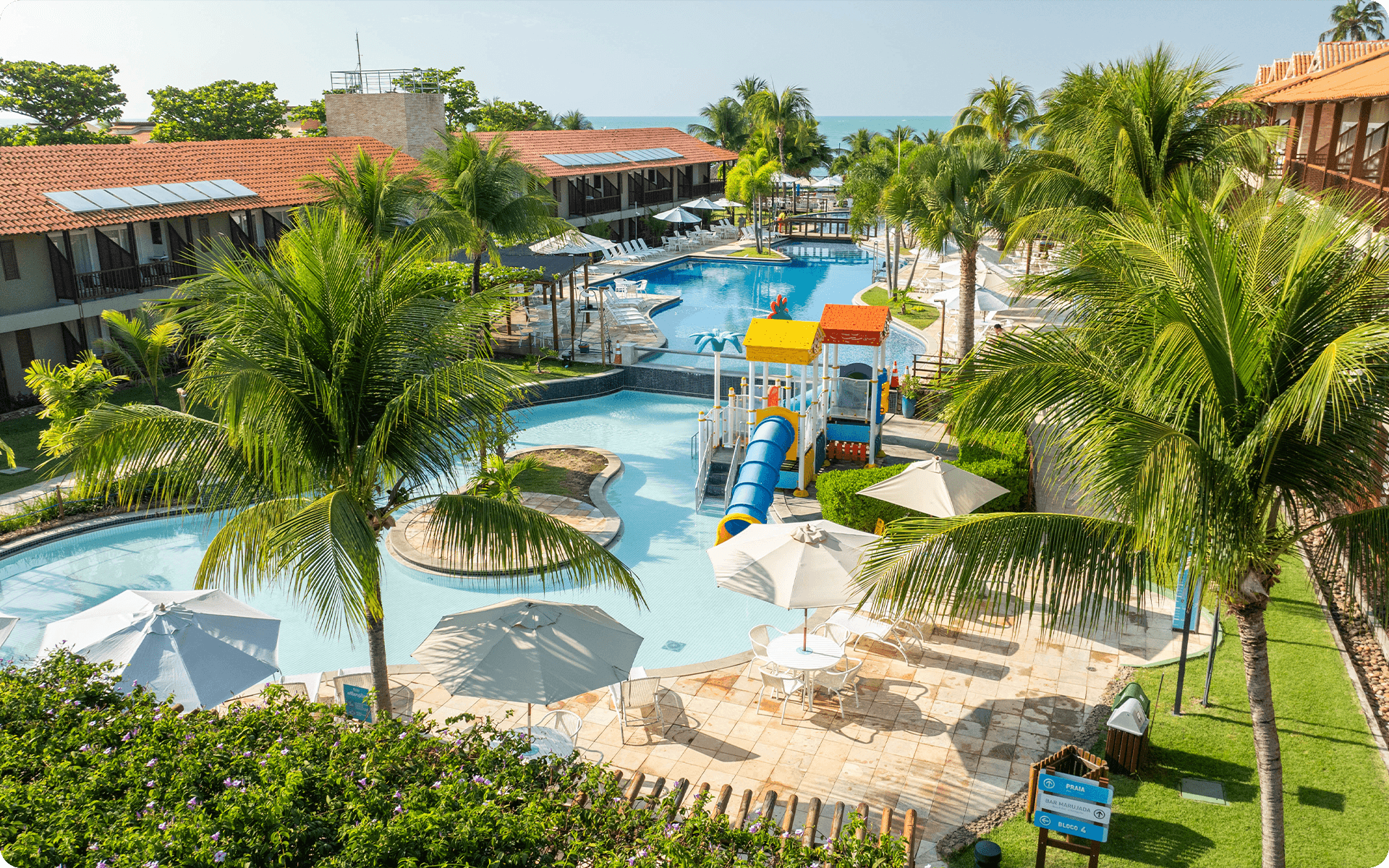 Complexo de pisicna do Salinas Maceió, com toboáguas, guarda-sóis brancos. Apartamentos ao redor e ao fundo pode se ver o mar e céu sem nuvens.