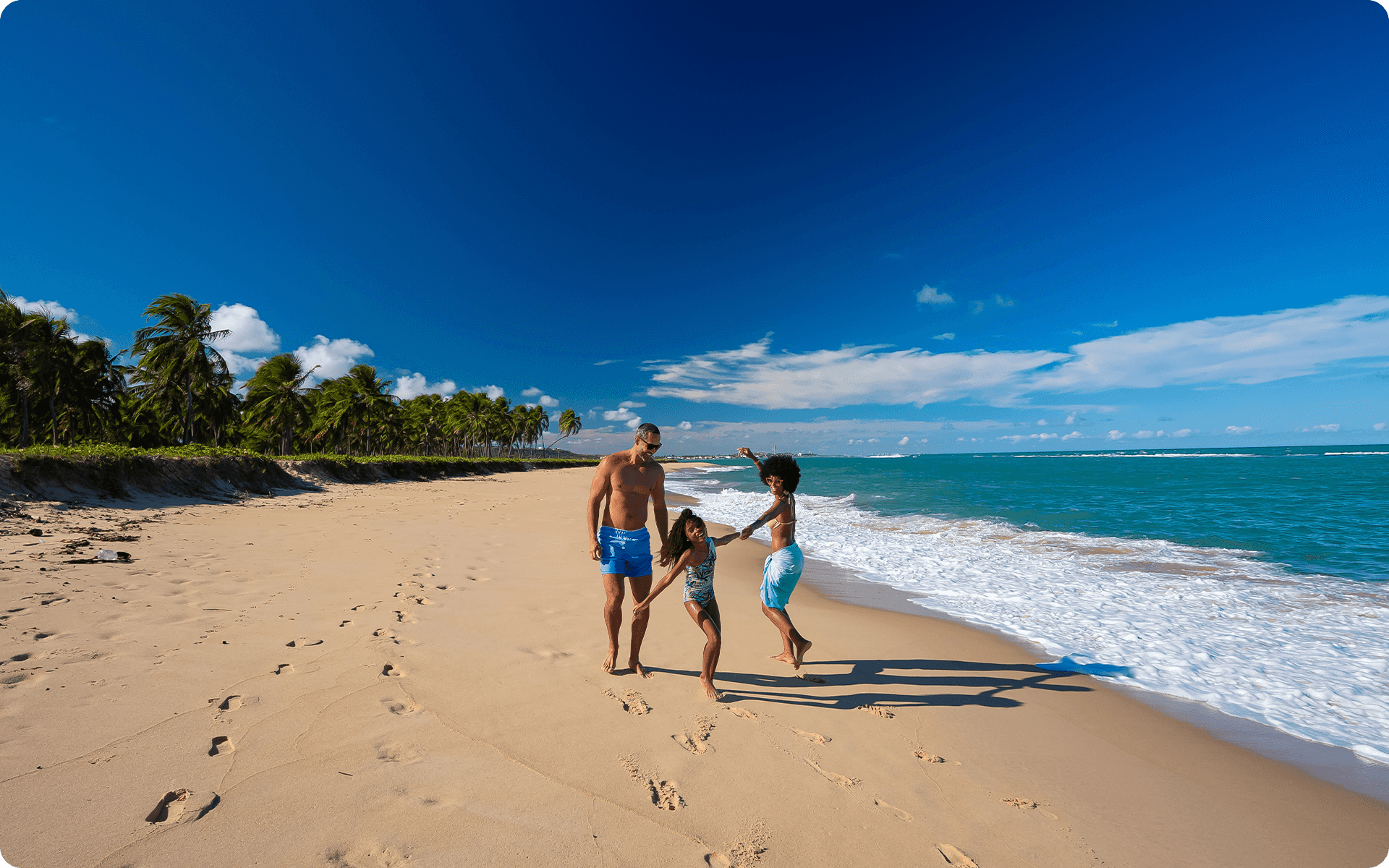 Na Praia do Gunga, o céu tem poucas nuvens, o mar está calmo. Uma família brinca na areia, com vários coqueiros ao fundo.