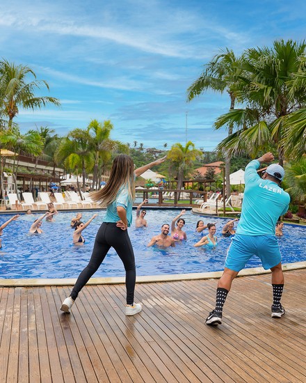 Recreação e hidroginástica na piscina do Salinas Maceió All Inclusive Resort Dois monitores da equipe de lazer em um deck de madeira orientando hóspedes em uma aula de hidroginástica dentro da piscina do resort, cercada por coqueiros e área de acomodações.
