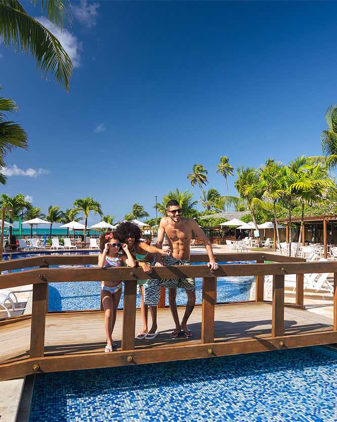 familia ponte piscina resort maceio Família sorrindo sobre uma pequena ponte de madeira que atravessa a piscina do resort. Eles estão em trajes de banho, sob um céu azul claro, rodeados por coqueiros, espreguiçadeiras e a estrutura do resort ao fundo.