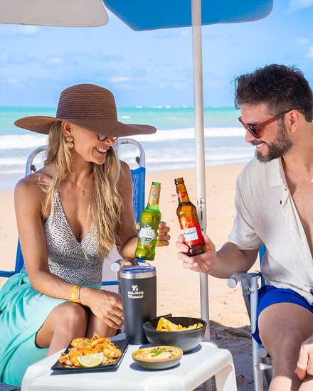 All Inclusive com petiscos e bebidas na praia em frente ao Salinas Maceió Casal sorrindo e brindando com garrafas de cerveja sob guarda-sol do Salinas Maceió na praia, com mesa servida de camarões grelhados e batatas fritas com o mar ao fundo.