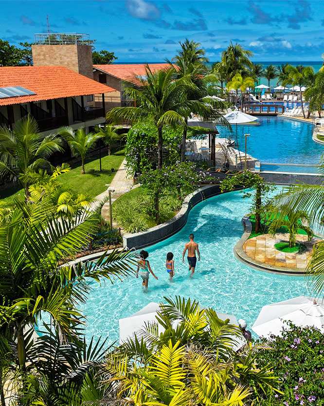 piscina familia resort Vista da grande piscina que se integra à paisagem do Resort Maceió, rodeada por vegetação tropical e coqueiros. Um casal com um filho caminha na água clara. Ao redor, espreguiçadeiras e guarda-sóis brancos, com os edifícios do resort Salinas Maceió ao fundo.