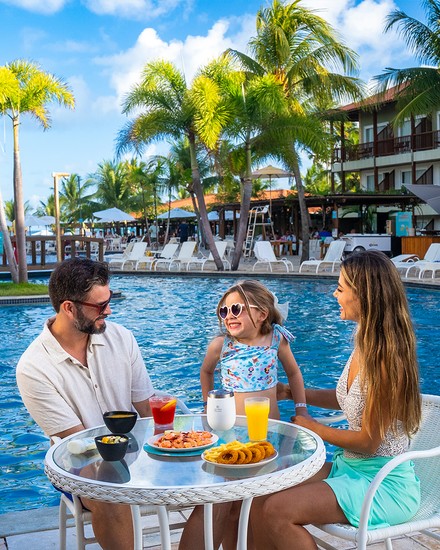 Família desfrutando de petiscos e bebidas à beira da piscina no Salinas Maceió Pai, mãe e filha pequena sentados em uma mesa à beira da piscina do resort. A mesa está servida com aperitivos e bebidas. Ao fundo, coqueiros, blocos de acomodação e o céu azul.