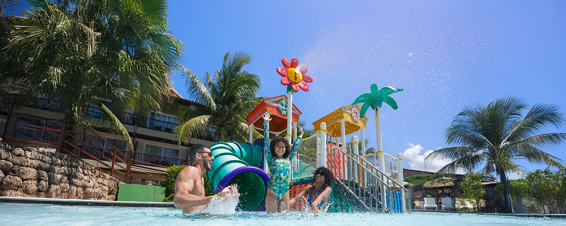 black friday salinas maceio Família se divertindo e brincando na área rasa da piscina infantil. A filha está animada com os braços erguidos. Ao fundo, o parque aquático colorido com toboáguas e elementos lúdicos, e coqueiros.