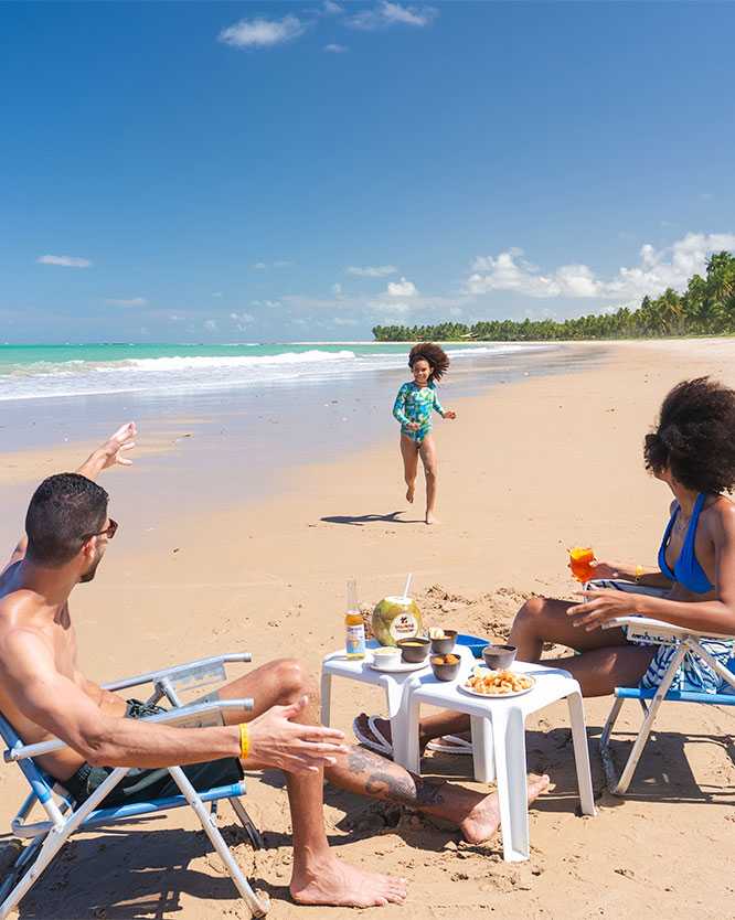 praia de ipioca alagoas Família relaxando em uma praia de areia clara. O casal está sentado em cadeiras de praia, com petiscos e bebidas em uma mesa. A filha corre e sorri na areia vinda da direção do mar. Um coqueiral e o mar azul-turquesa estão no fundo.