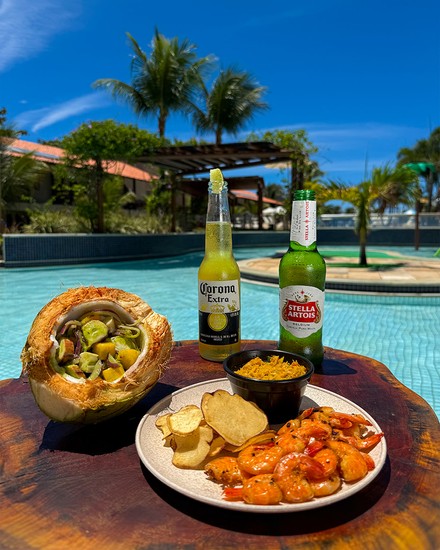 Gastronomia e aperitivos à beira da piscina no Salinas Maceió All Inclusive Resort Mesa de madeira à beira da piscina com prato de camarões grelhados e batatas, petisco servido no coco seco e duas garrafas de cerveja geladas sob dia ensolarado.