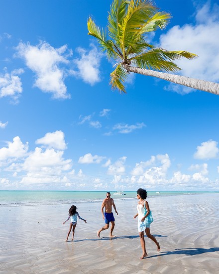 um destino encantador que te convida a ser feliz Família brincando em uma extensa faixa de areia da praia de Ipioca. O mar está com a maré baixa e existe um grande coqueiro próximo a eles. O dia está ensolarado com céu azul com poucas nuvens.