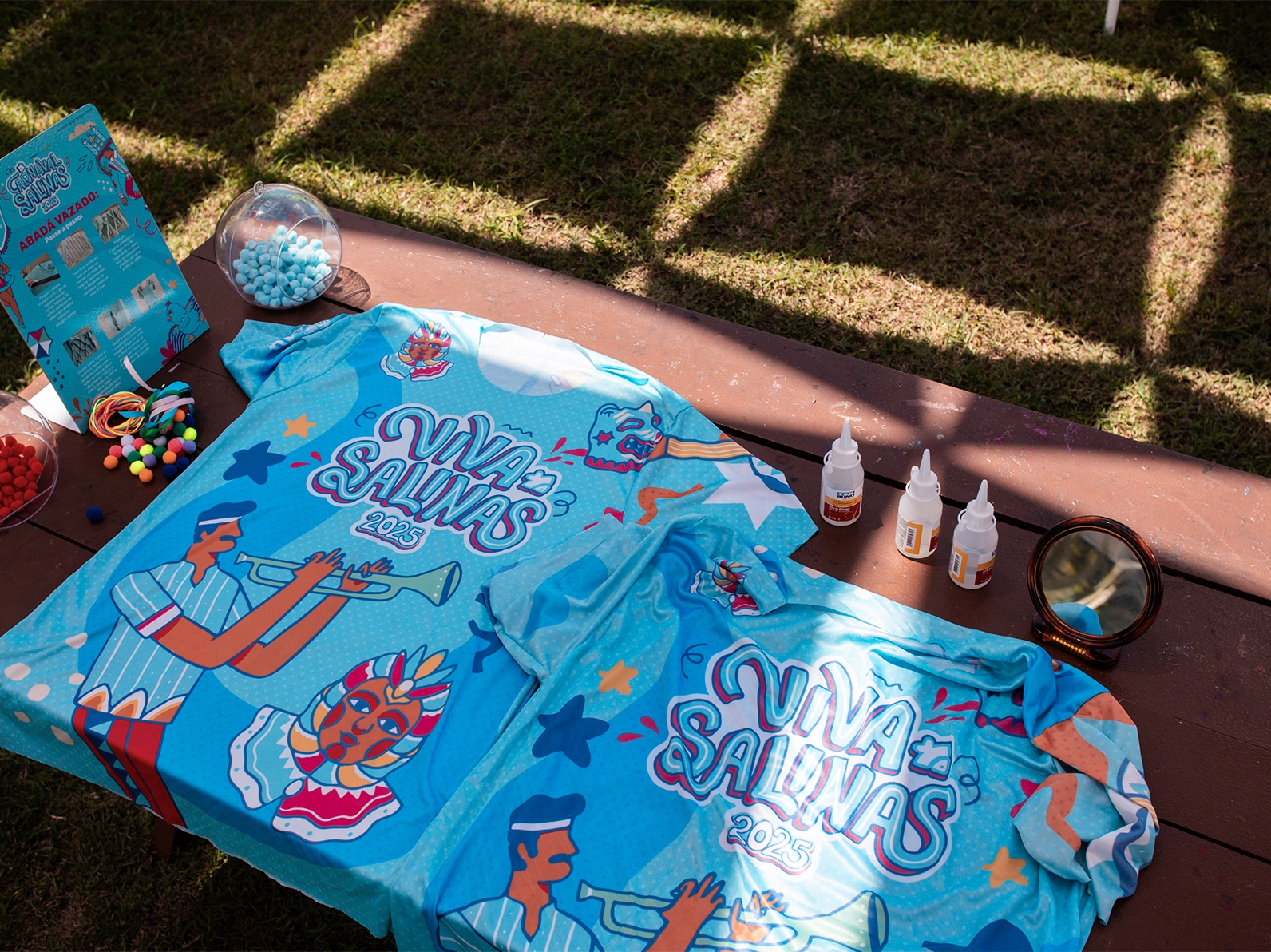 Mesa de madeira na grama, sobre ela tem os abadás com a identidade visual do bloco, de cor azul com detalhes coloridos, e ao redor tem materiais pra customizá-las..