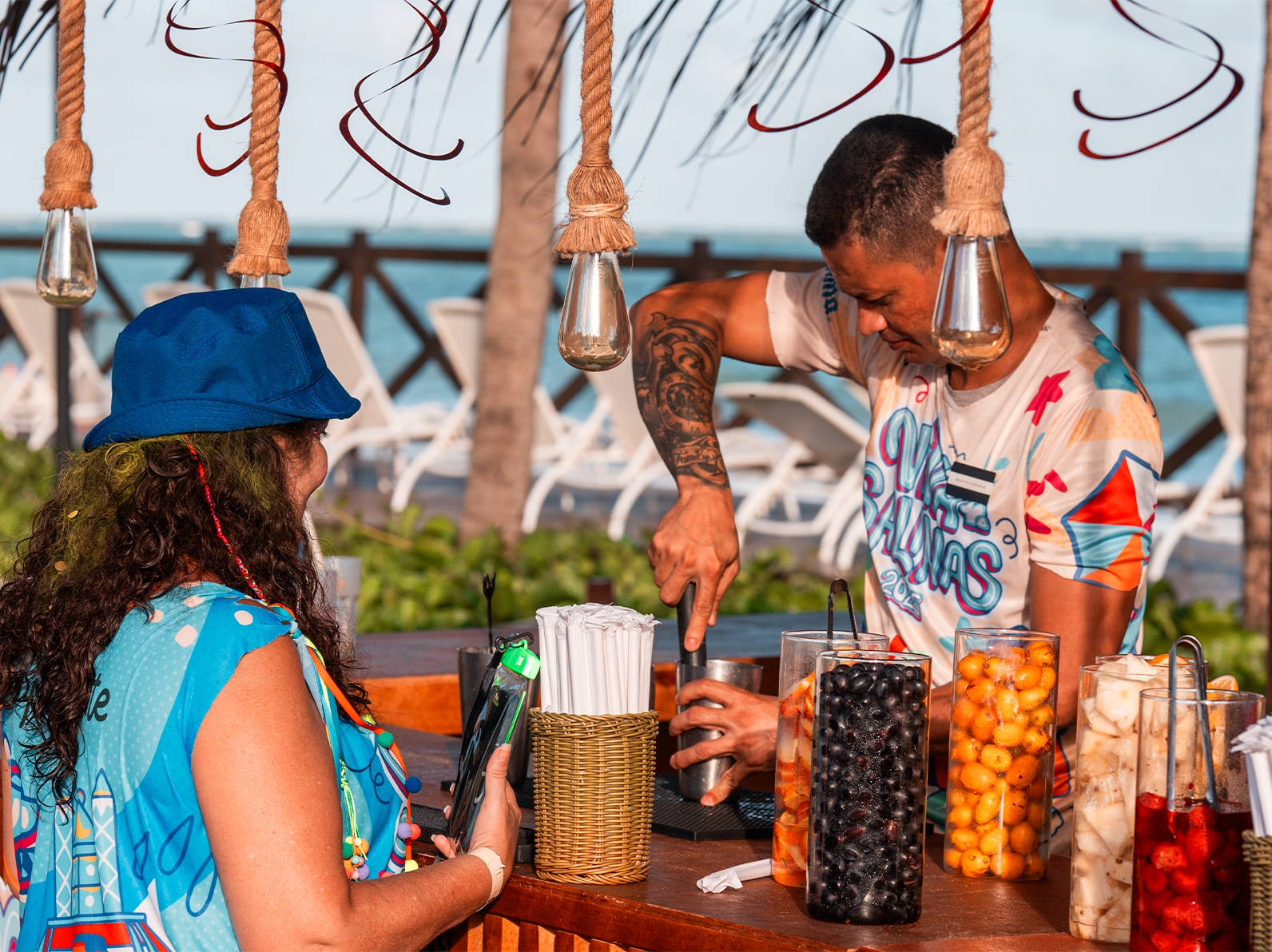 Bar externo pro carnaval Viva Salinas. Balcão de madeira, com pendentes de luz pendurados, o bartender prepara um drink pra uma hóspede que está com o abadá, está com dia ensolarado e há várias frutas no balcão.