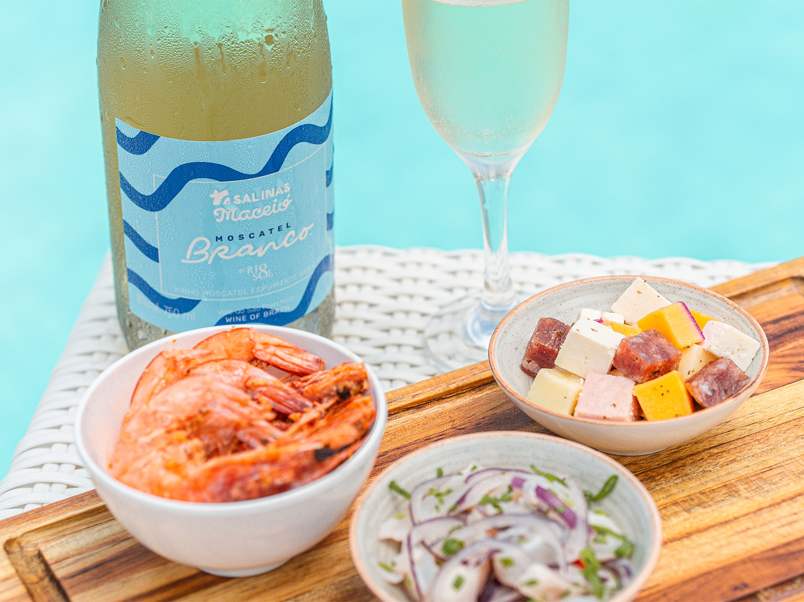 Garrafa de moscatel branco da marca RioSol com taça ao lado, tem petiscos, uma porção de camarão, queijos e ceviche em frente a piscina.
