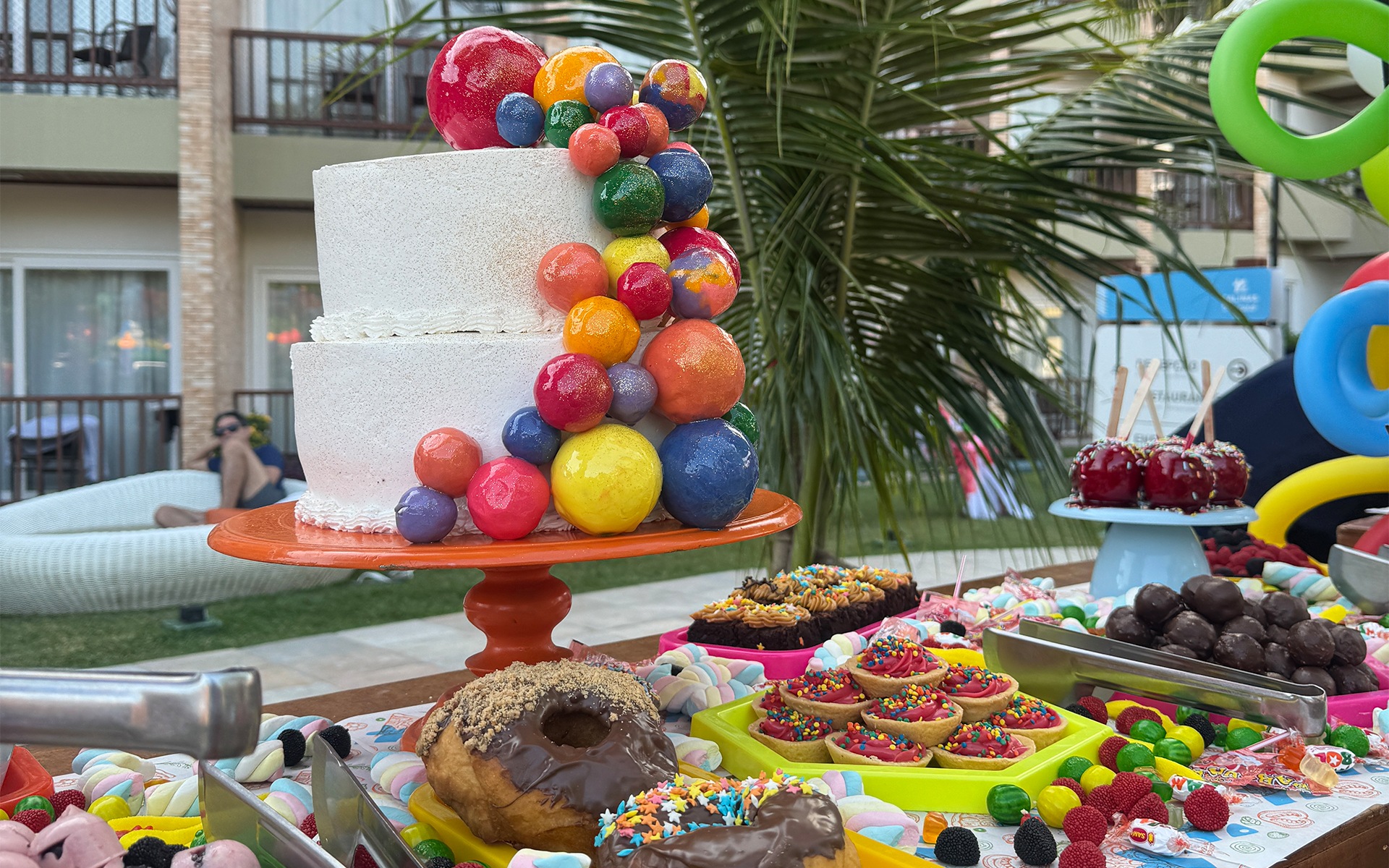 Mesa de doces coloridos com bolo decorado e guloseimas em área externa do Salinas Maceió, preparada para programação infantil durante o dia das crianças.