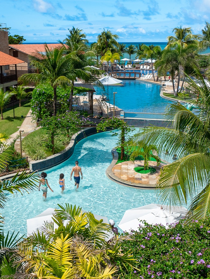 Área de lazer do Salinas Maceió, com uma pequena ilha no meio da piscina. Estrutura dos quartos ao redor e a praia ao fundo com céu sem nuvens.
