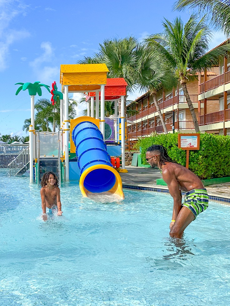 Família se divertindo na piscina infantil do Salinas Maceió, com brinquedos aquáticos coloridos, crianças brincando e estrutura ao fundo.