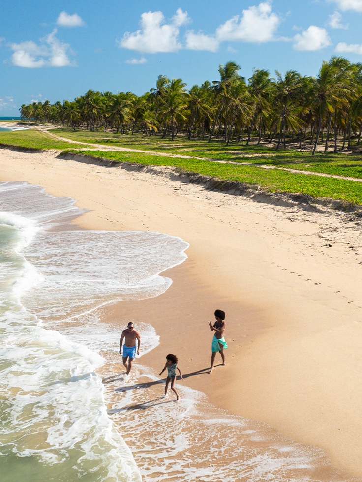 Família caminhando à beira-mar na Praia do Gunga, em Alagoas, com marzão esverdeado e coqueiros.