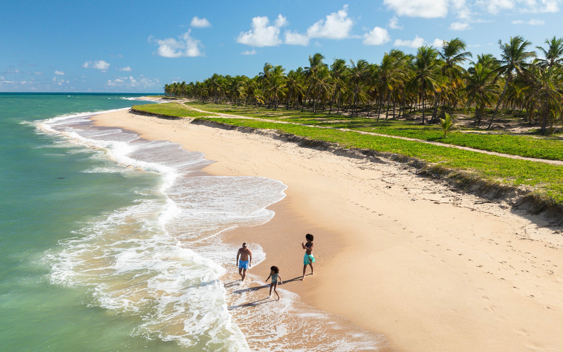 Família caminhando à beira-mar na Praia do Gunga, em Alagoas, com marzão esverdeado e coqueiros.