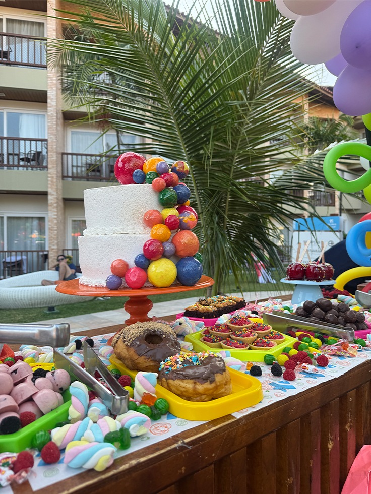 Mesa de doces coloridos com bolo decorado e guloseimas em área externa do Salinas Maceió, preparada para programação infantil durante o dia das crianças.