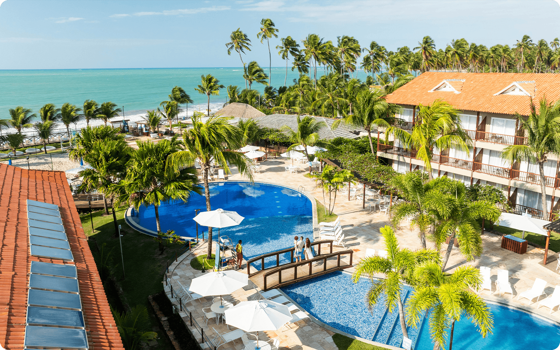Área de lazer do Salinas Maceió, com ponte cruzando a piscina principal. Estrutura dos quartos ao redor e a praia ao fundo com céu sem nuvens.