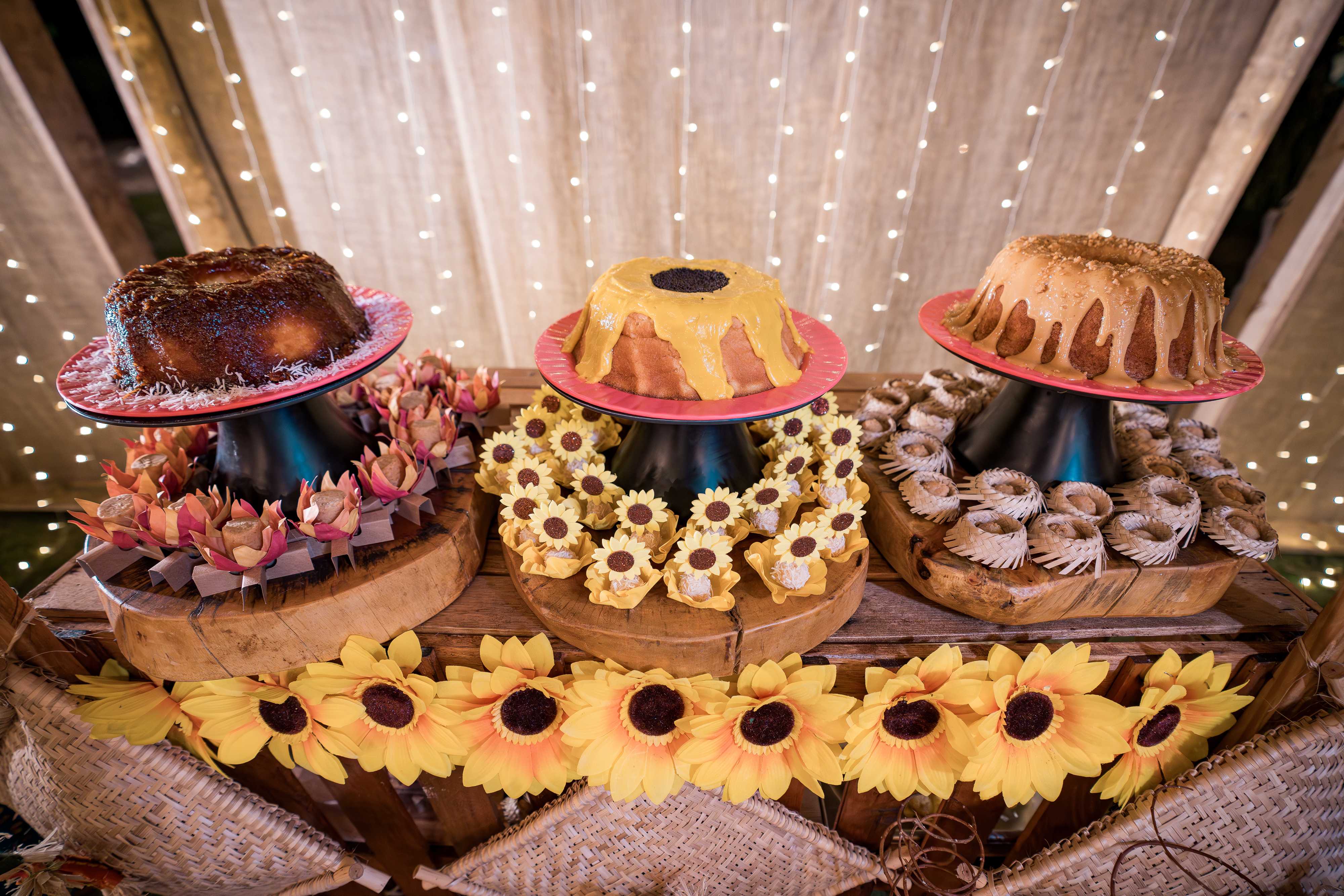 Mesa decorada com girassóis e luzes, exibindo bolos e docinhos típicos de festa junina.