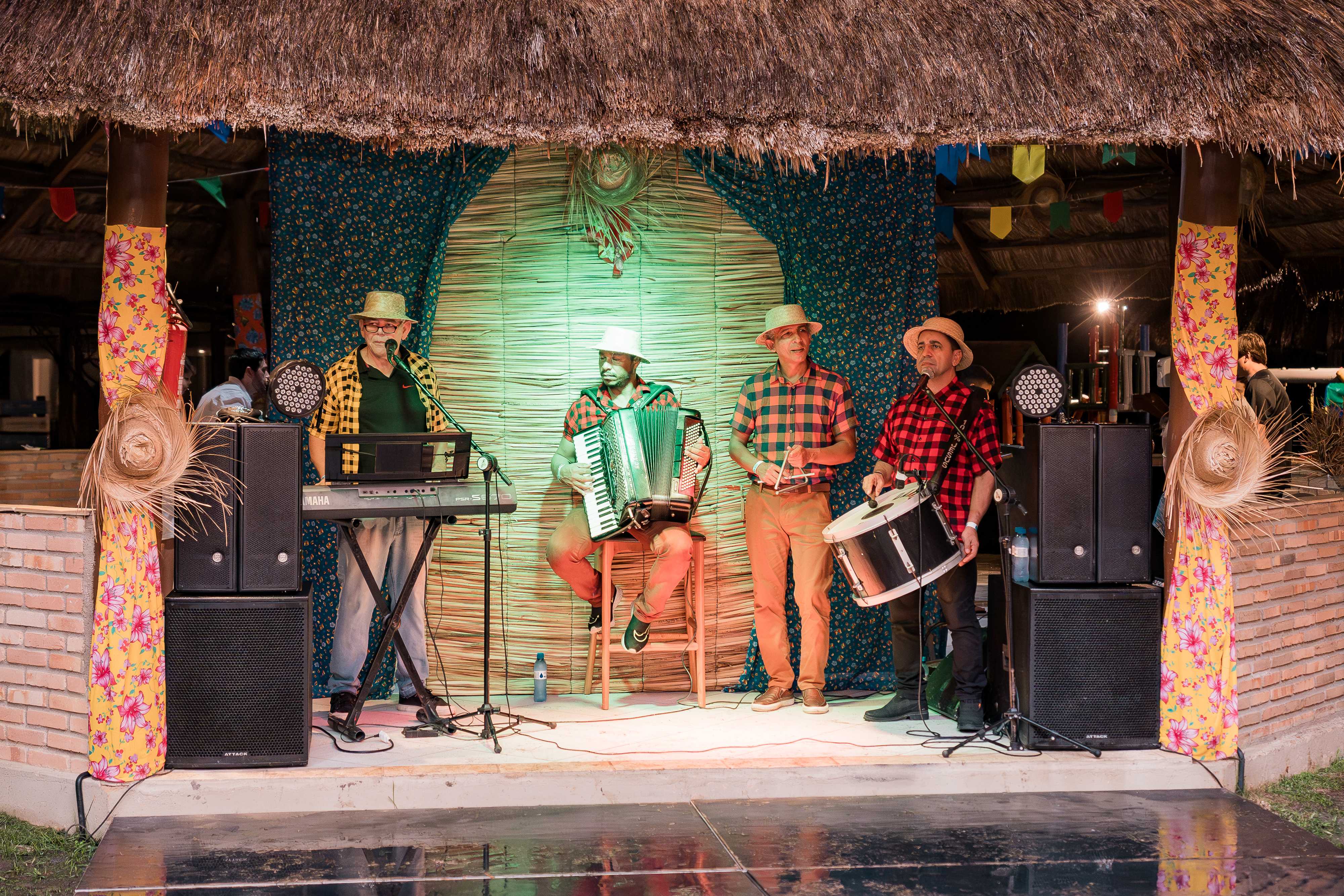 Banda se apresentando em uma festa junina, com músicos tocando teclado, sanfona e percussão, todos vestidos com trajes típicos.