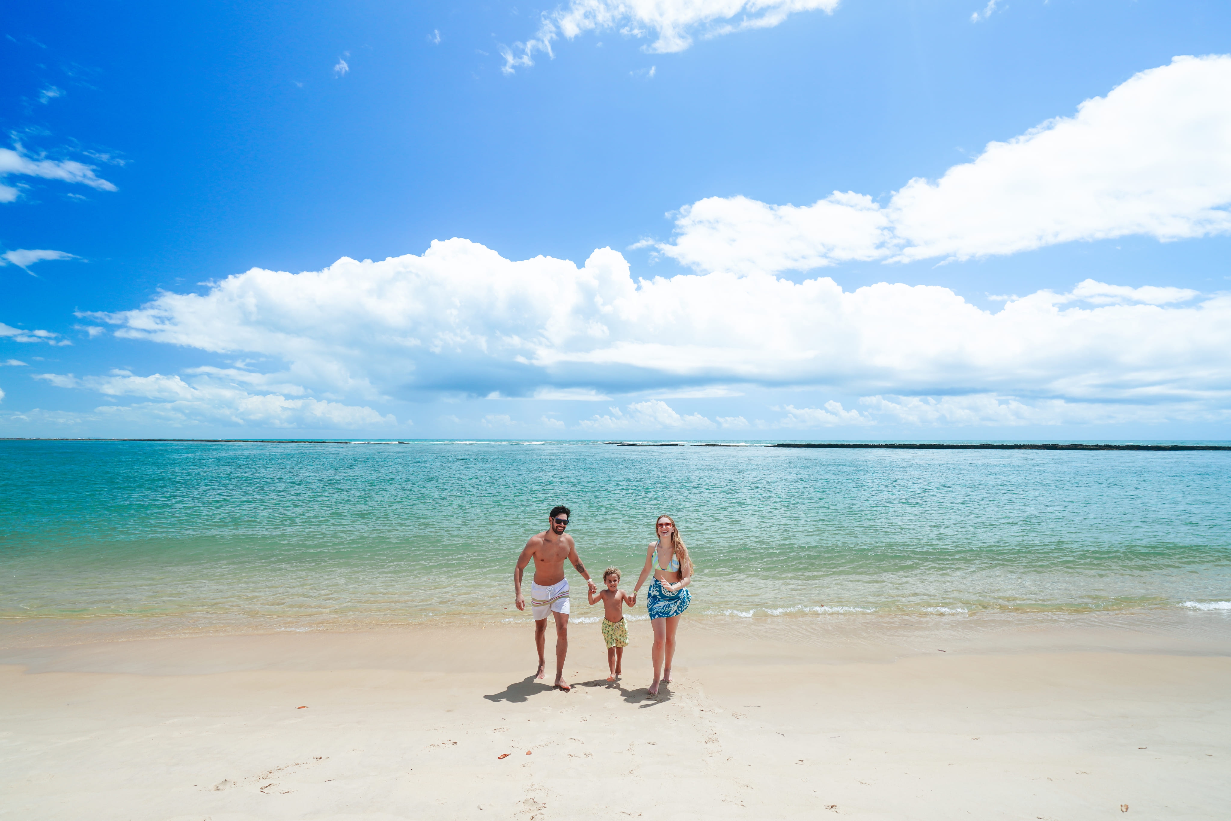 Família caminha sob areia branca à beira-mar da praia de Ipioca, em Maceió.