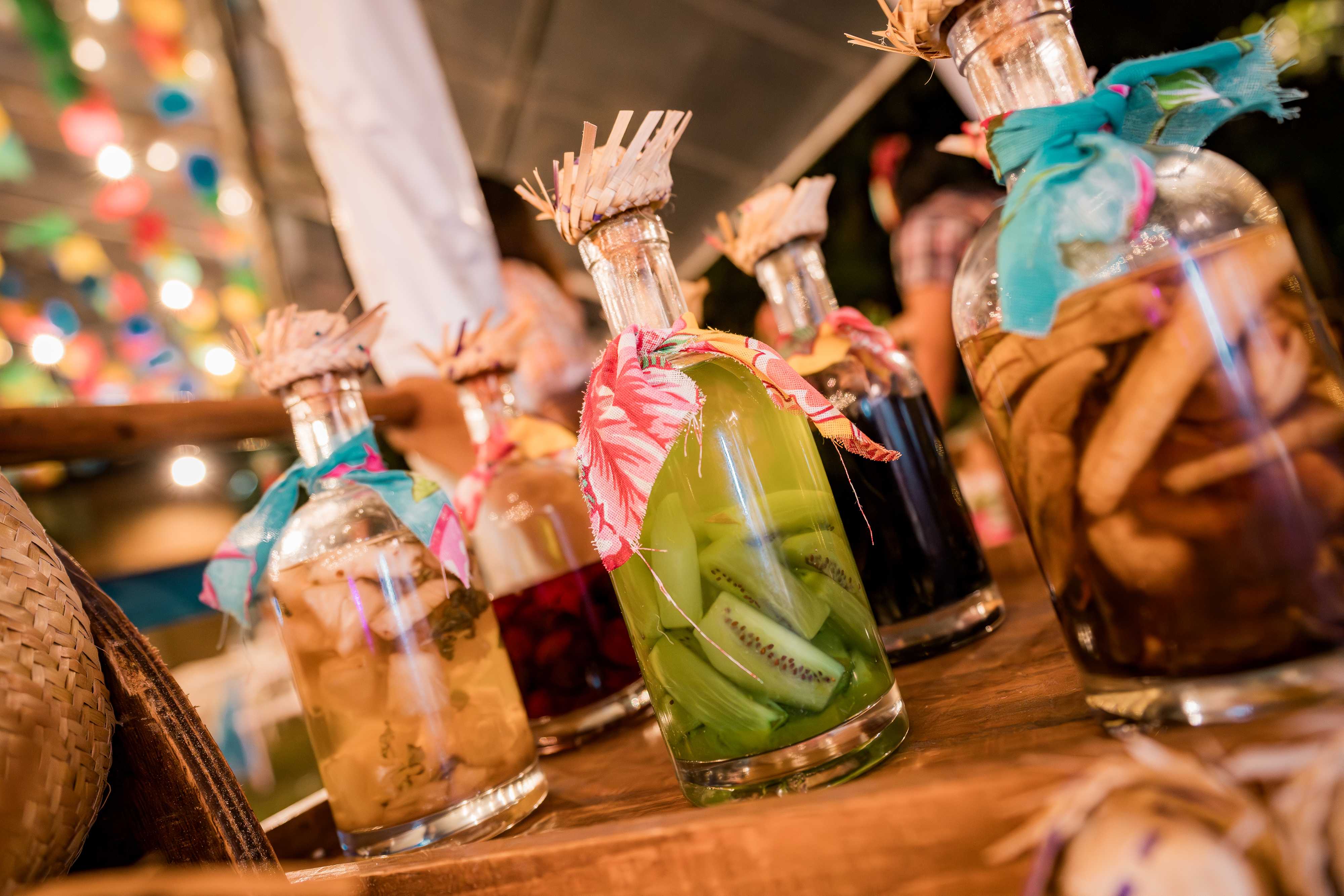 Garrafas de bebidas artesanais decoradas com panos coloridos em uma festa junina, com frutas e especiarias dentro das garrafas.