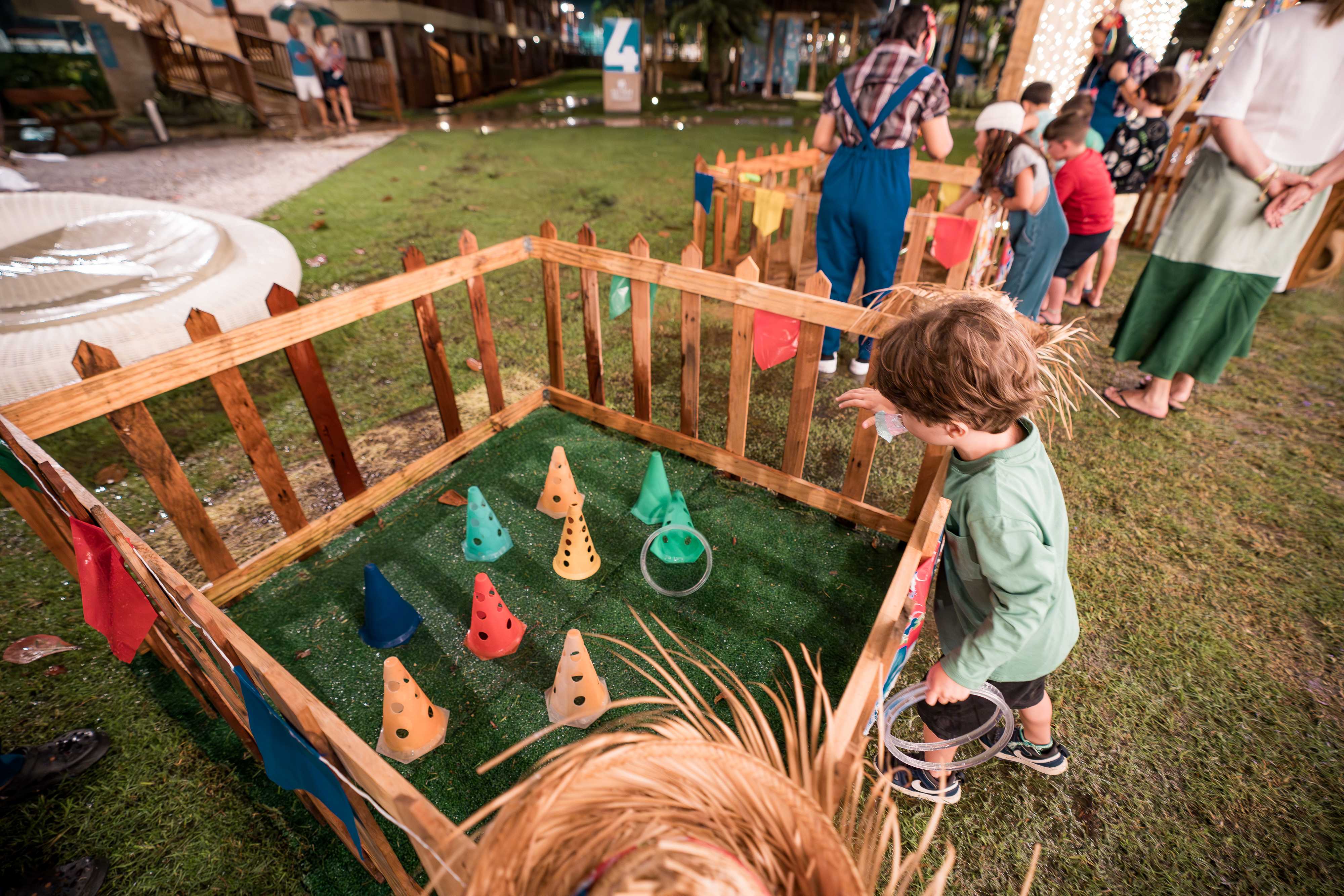 Criança brincando de argolas em uma festa junina, cercada por uma cerca de madeira com bandeirinhas coloridas.