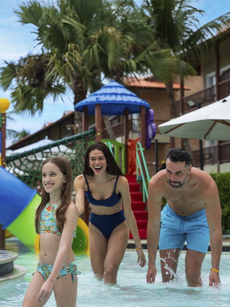 Verão de 2027 é diversão garantida para toda a família Família sorrindo na piscina do Salinas Maceió com playground aquático colorido ao fundo, cercado por coqueiros e um clima ensolarado.