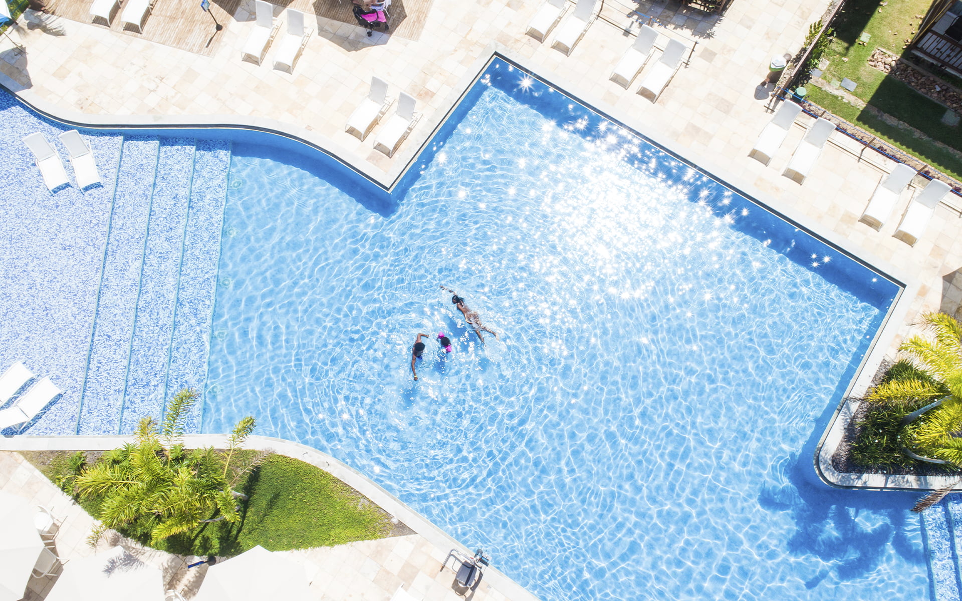 A piscina que reflete o brilho das suas próximas férias Vista aérea da piscina do Salinas Maceió com pessoas nadando, um clima ensolarado e ambiente cercado por espreguiçadeiras e jardim verde.