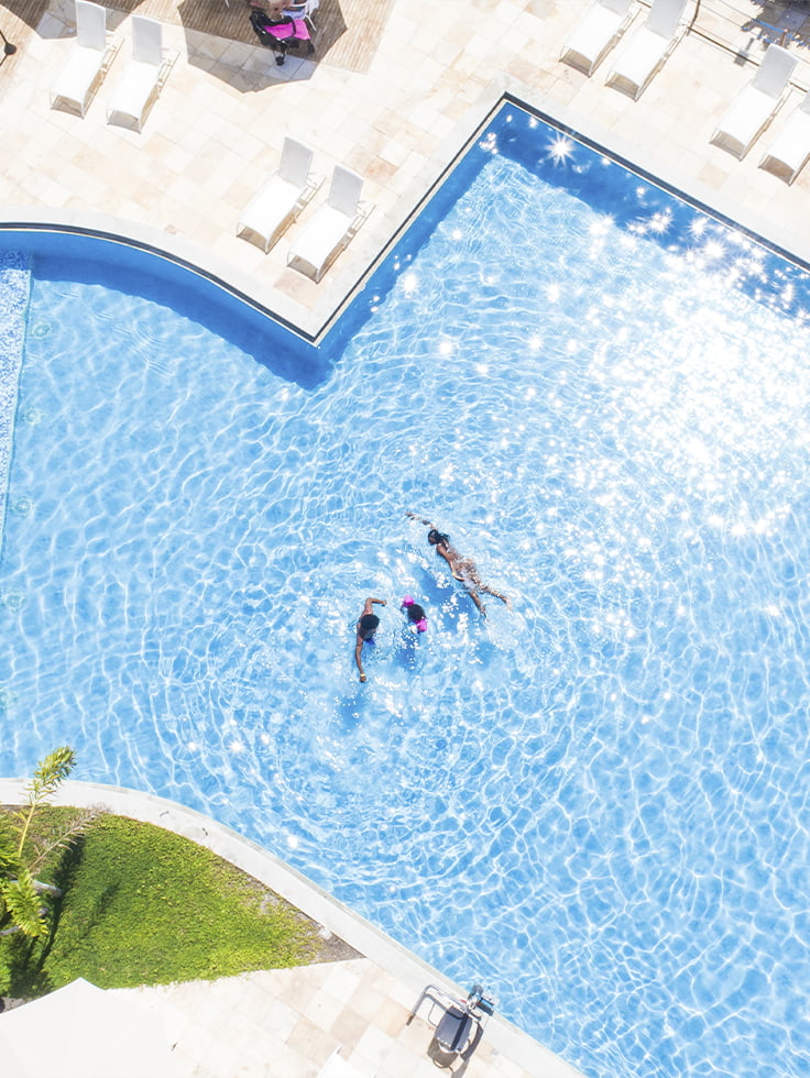 A piscina que reflete o brilho das suas próximas férias Vista aérea da piscina do Salinas Maceió com pessoas nadando, um clima ensolarado e ambiente cercado por espreguiçadeiras e jardim verde.