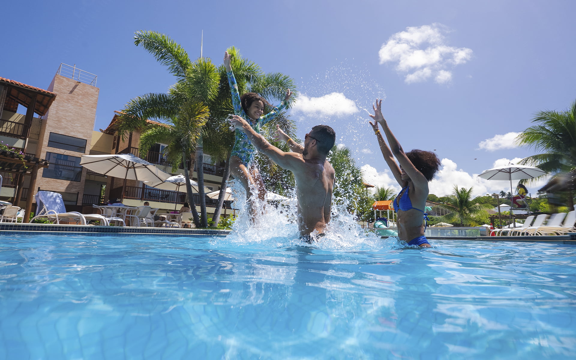 Um mergulho de pura energia em família Pai segurando filha ao pular na piscina do Salinas Maceió, com mãe comemorando ao lado, respingos de água e estrutura dos quartos do resort ao fundo.