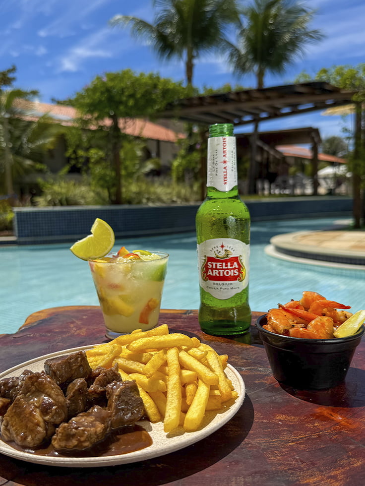 Uma estrutura completa até na hora da fome Mesa à beira da piscina do Salinas Maceió com caipirinha, cerveja Stella Artois, batata frita, camarão e petisco de carne, cercada por área de lazer e coqueiros.