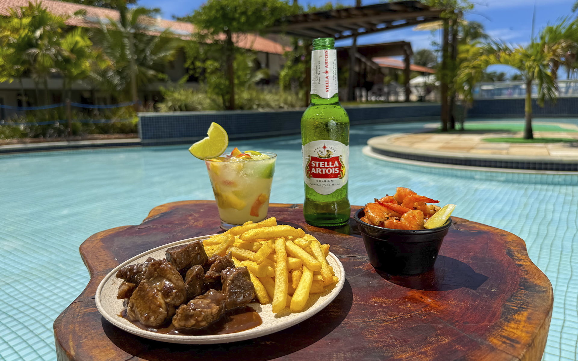 Uma estrutura completa até na hora da fome Mesa à beira da piscina do Salinas Maceió com caipirinha, cerveja Stella Artois, batata frita, camarão e petisco de carne, cercada por área de lazer e coqueiros.