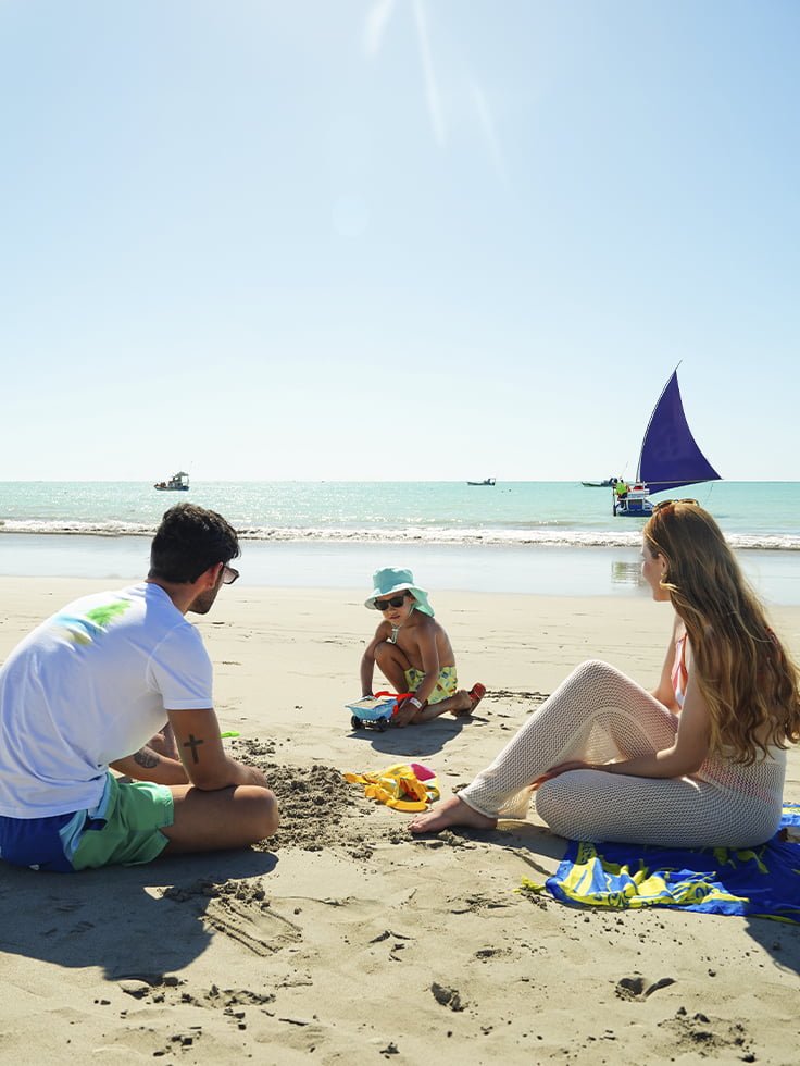Memórias pra guardar pra sempre na Praia de Pajuçara Família sentada na areia da Praia de Pajuçara em Maceió, com jangada de vela e barcos ao fundo em dia de céu claro.