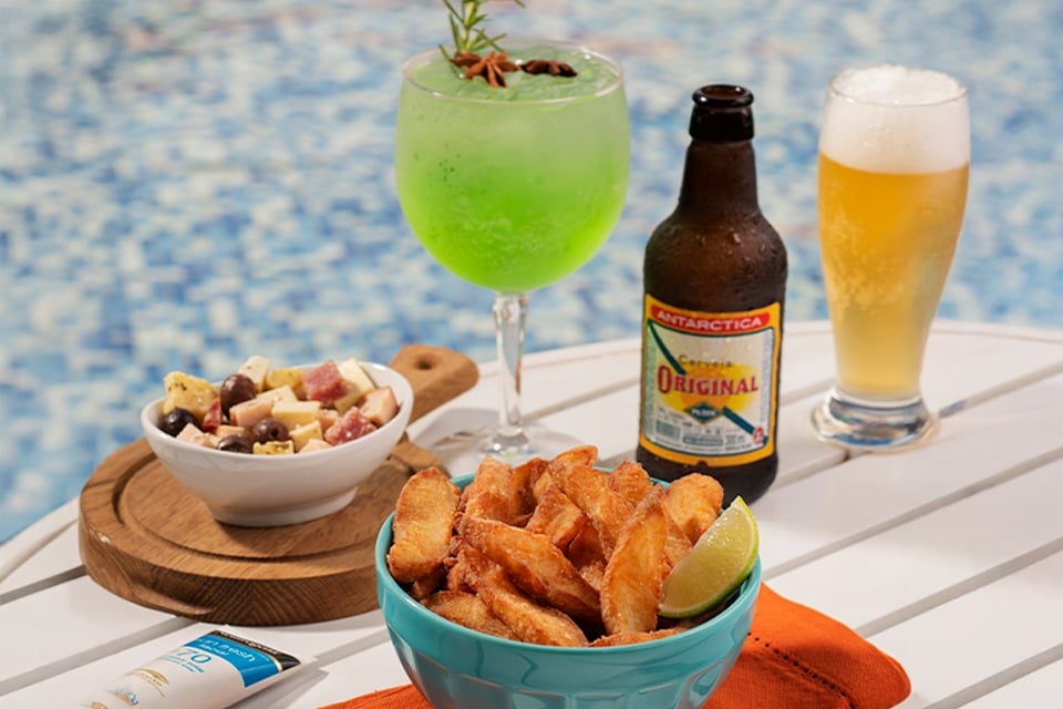 Cerveja long neck Original, taça de drink e bowls de petiscos sobre mesa à beira da piscina do Salinas Maceió.