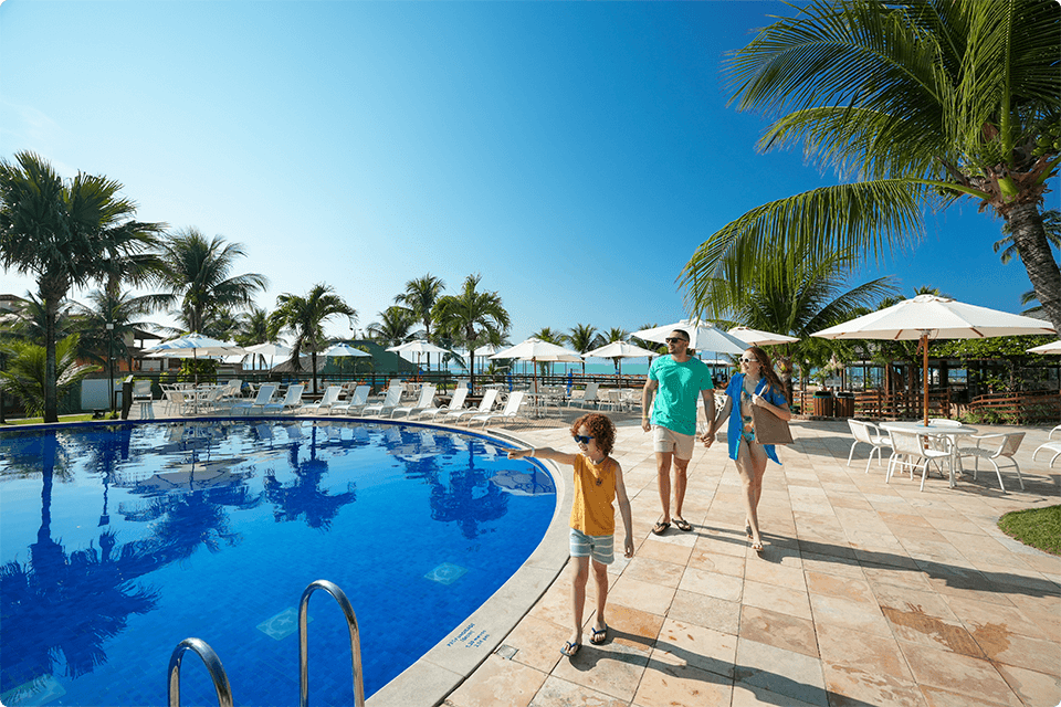 Família caminhando na beira da piscina do resort, com coqueiros, cadeiras de sol brancas ao fundo. Um bar do lado direito e um céu azul sem nuvem.